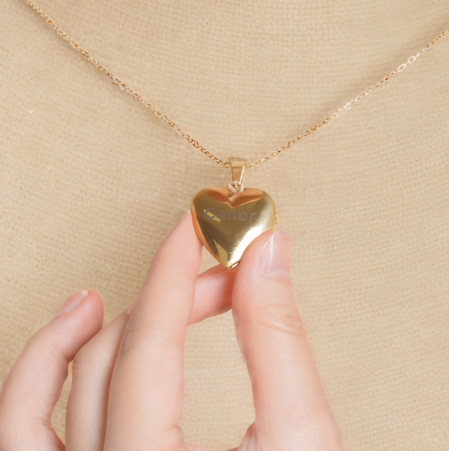 Close up of gold Heart Locket Necklaces worn on a delicate chain with hand touching pendant on beige top