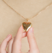 Close up of gold Heart Locket Necklaces worn on a delicate chain with hand touching pendant on beige top